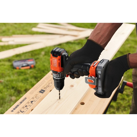 BLACK+DECKER cordless drill in use, drilling into a wooden plank outdoors with gloved hands.