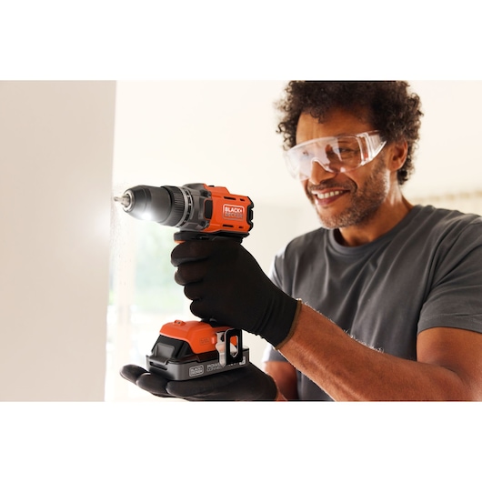 Person uses BLACK+DECKER cordless drill on a wall, wearing gloves and safety goggles indoors.