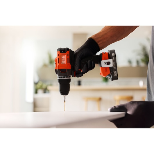 A BLACK+DECKER cordless drill is used to drive a screw into a white surface in a modern indoor setting.