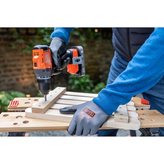 Person uses BLACK+DECKER cordless drill to screw wood pieces with gloves on a workbench outdoors.