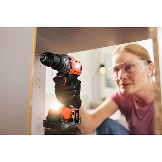 Person uses a BLACK+DECKER cordless drill to assemble furniture, wearing safety glasses and gloves.