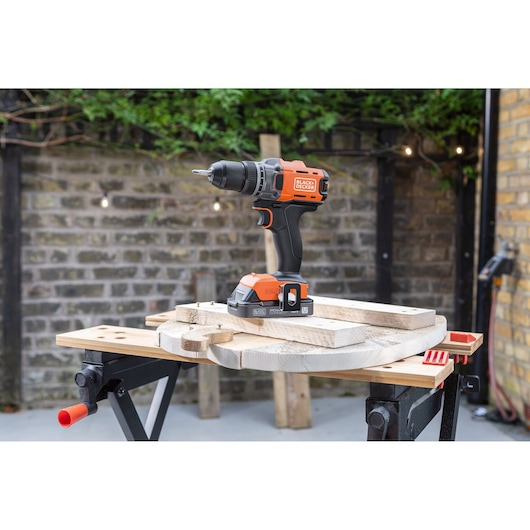 BLACK+DECKER cordless drill stands on a wooden board outdoors, set on a workbench with tools nearby.