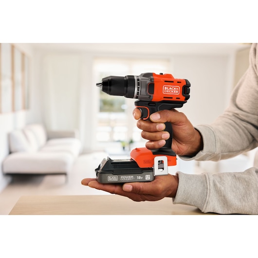 A BLACK+DECKER cordless drill held in hands, shown indoors with a blurred sofa in the background.