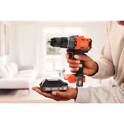 Person holding a BLACK+DECKER cordless drill and battery, shown in a bright living room setting.