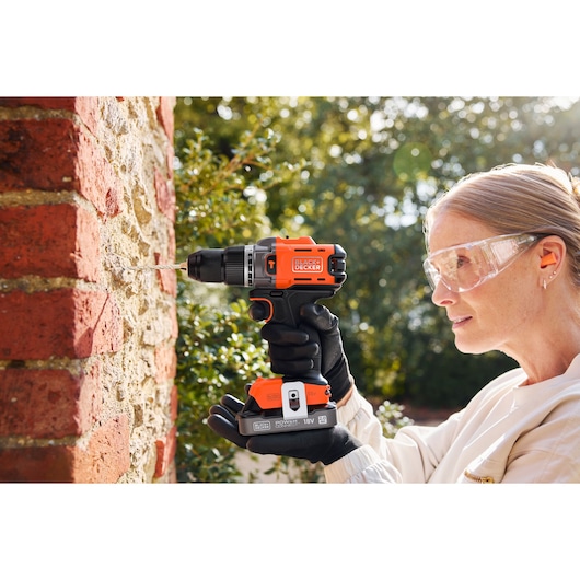 Person drills into outdoor brick wall using a BLACK+DECKER cordless drill, wearing safety glasses and gloves.