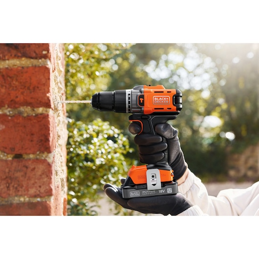 BLACK+DECKER cordless drill in use, drilling into a brick wall outdoors, held by a gloved hand.