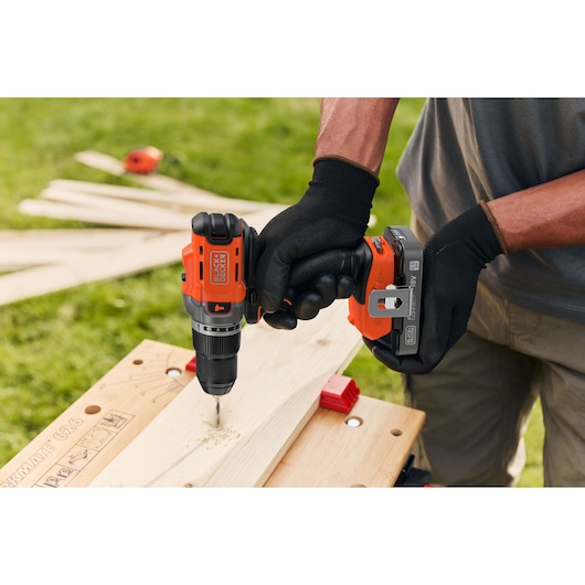 BLACK+DECKER cordless drill being used to drill a hole in wood outdoors by gloved hands.