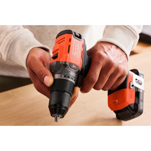 Close-up of hands holding a BLACK+DECKER cordless drill with an orange design on a light wooden surface.