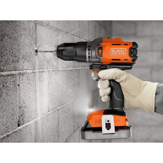 A BLACK+DECKER cordless drill in use, drilling into a concrete wall, held by a gloved hand.