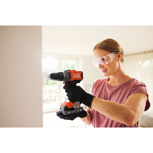 Person drilling into a wall with a BLACK+DECKER cordless drill, wearing safety glasses and gloves indoors.