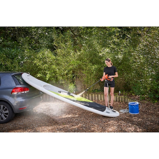 A person using a BLACK+DECKER portable pressure washer to clean a paddleboard, which is resting on the back of a car, outdoors. A bucket of water is placed nearby and the area is surrounded by greenery.