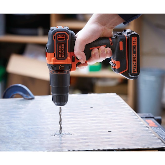 A person using a BLACK+DECKER cordless drill with an 18V lithium battery to drill into a metal surface.