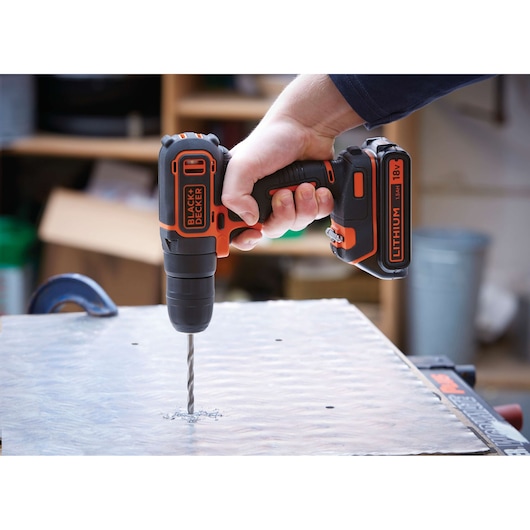 A BLACK+DECKER 18V lithium cordless drill being used to drill into a metal sheet in a workshop setting.
