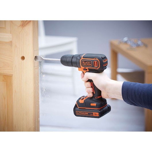 A person is using a BLACK+DECKER 18V lithium cordless drill to drill into a wooden door. The environment appears to be indoors with furniture in the background.