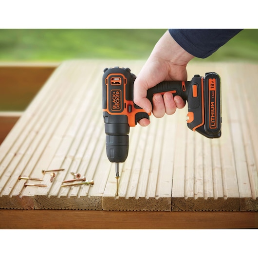 A person is using a BLACK+DECKER cordless drill to screw a bolt into wooden planks. Several screws are resting nearby on the wood. The drill is branded with 'BLACK+DECKER' and 'Lithium 18V'.