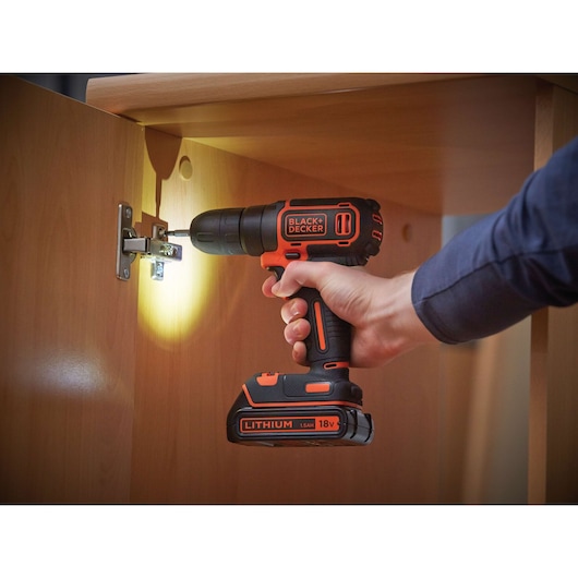 A person using a BLACK+DECKER 18V lithium cordless drill to install a hinge on a wooden cabinet door. The drill has an LED light illuminating the work area.