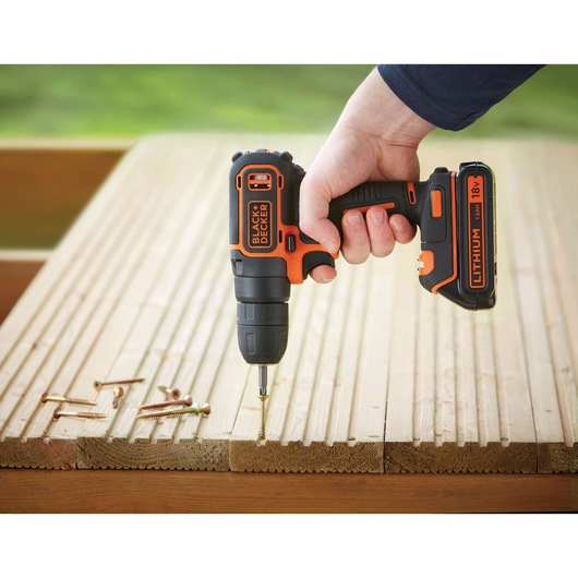 A hand holding a BLACK+DECKER cordless drill, driving a screw into a wooden surface. Several screws are also lying on the wood.