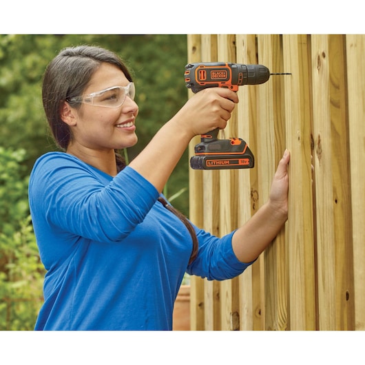 A person in a blue shirt is using a BLACK+DECKER BDCDC18 cordless drill to drill into a wooden fence outdoors. The person's face is blurred.