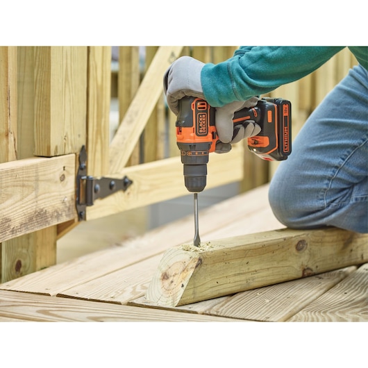 A person wearing gloves using a BLACK+DECKER cordless drill to drill into a wooden plank outdoors.