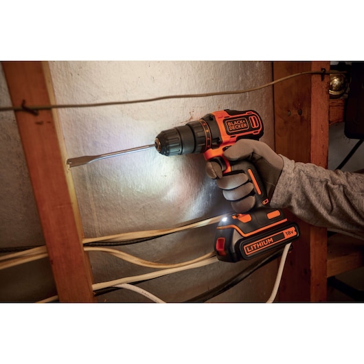 A person wearing gloves is using a BLACK+DECKER 18V cordless drill with a lithium battery to drill a hole in a wall, with electrical wires visible nearby.