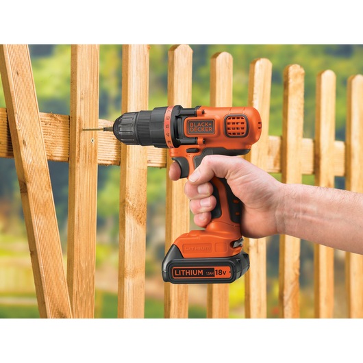 A person using an orange and black cordless BLACK+DECKER power drill to screw into a wooden fence outdoors.