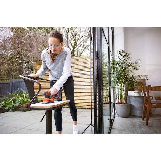 A person using a BLACK+DECKER cordless sander on a chair outdoors, near a patio and an open glass door, with plants and furniture visible inside.