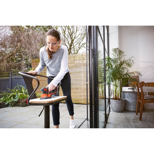 A person is sanding a wooden chair outdoors using a BLACK+DECKER BDCDS18 cordless sander. The chair is placed near a sliding glass door with a view of a modern interior and plants.