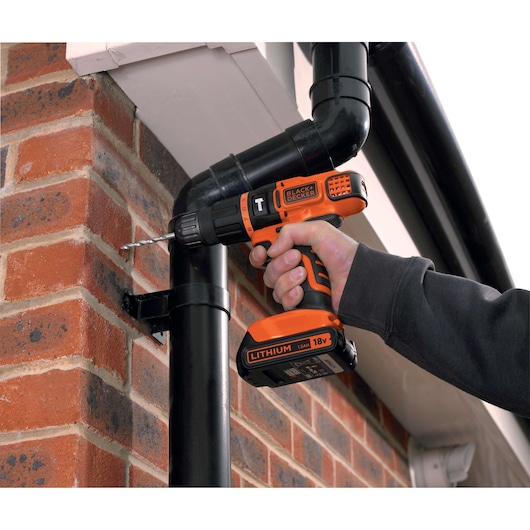 A person using a BLACK+DECKER cordless drill to drill into a brick wall near a black pipe outdoors.