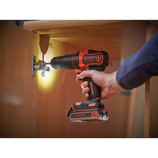 A person using a BLACK+DECKER 18V cordless drill with built-in light to install a hinge inside a wooden cabinet.