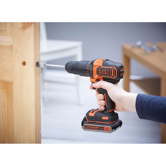 A person using a BLACK+DECKER 18V cordless drill to drill a hole into a wooden door. The drill features an orange and black design and is equipped with a lithium battery. Sawdust is visible as the drill bit cuts into the wood.