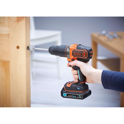 A person uses a BLACK+DECKER 18V cordless drill to drill a hole in a wooden door. The drill has orange and black accents and is labeled with 'smart tech' and '18V Lithium'.