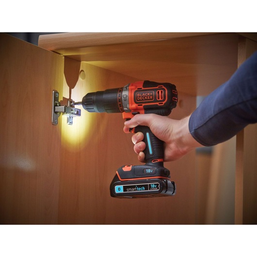 A person using a BLACK+DECKER 18V cordless drill with smart tech, drilling a screw into the hinge of a wooden cabinet door. The drill has an LED light illuminating the work area.