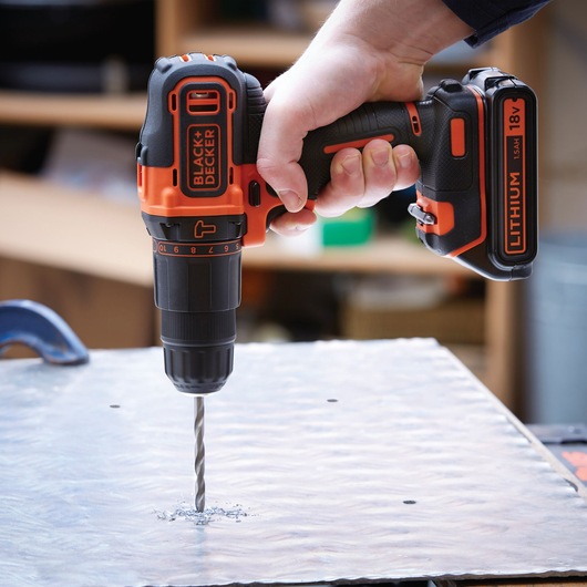 A person is using a BLACK+DECKER cordless power drill with an 18V lithium battery to drill a hole in a metal sheet.