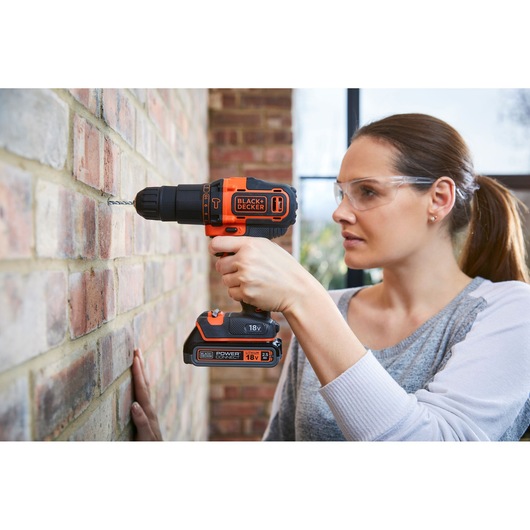 A person uses a BLACK+DECKER BDCHD18K cordless drill to drill into a brick wall. The person's face is obscured for privacy. The drill is orange and black and labeled with brand and battery information.