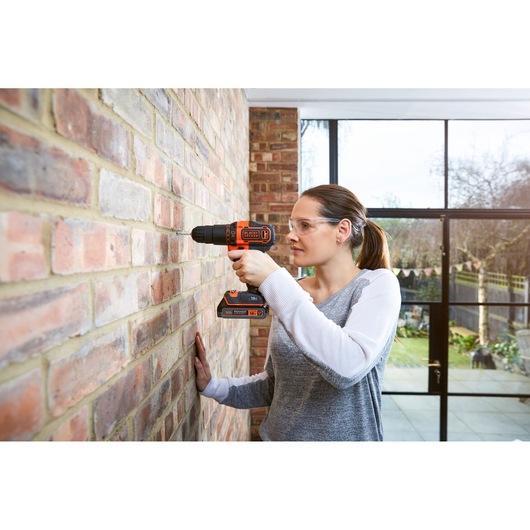 A person drills into a brick wall using a BLACK+DECKER BDCHD18K cordless power drill indoors. Their face is blurred for privacy.