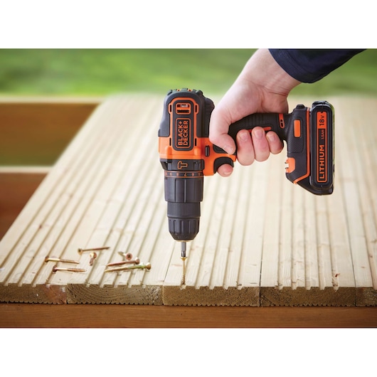 A BLACK+DECKER cordless drill, model BDCHD18, being used to drive a screw into wooden planks. Several screws are lying on the wood surface, and the drill is powered by an 18V lithium battery.