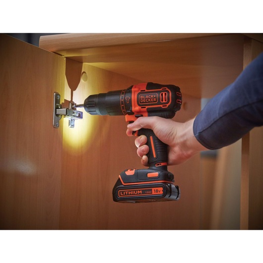 A BLACK+DECKER 18V cordless drill with lithium battery being used to screw a hinge onto a wooden cabinet. The drill is held by a person and a built-in light illuminates the hinge area.