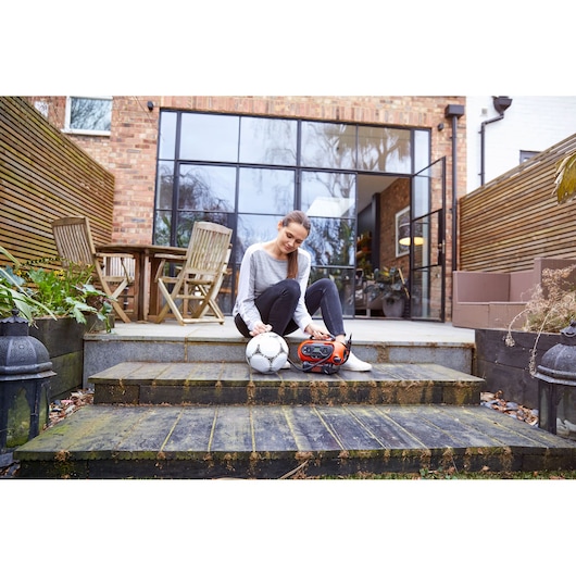 A person sitting on outdoor steps next to a soccer ball and a BLACK+DECKER portable inflator (model BDCINF18N), with a patio, wooden furniture, and glass doors in the background.