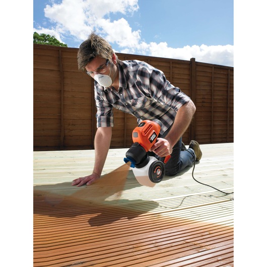 Person using BLACKDECKER BDPS200 paint sprayer on outdoor wooden deck, fence in background, low angle.