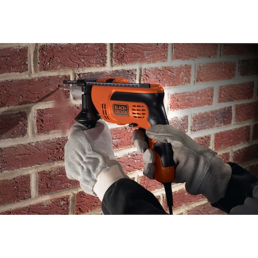 A person wearing protective gloves uses a BLACK+DECKER electric drill to drill into a brick wall.
