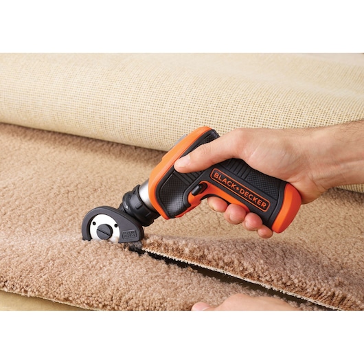 A person is using a BLACK+DECKER electric cutting tool to cut through a piece of beige carpet. The tool has a black and orange handle and is designed for precise cutting.