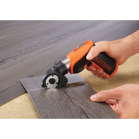 A person is using a BLACK+DECKER cordless rotary cutter to cut a piece of laminate flooring. The tool has an orange and black handle and is designed for precision cutting.