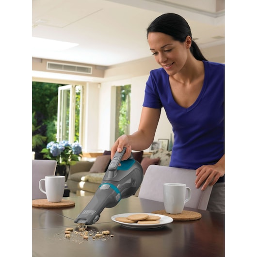 A person using a BLACK+DECKER handheld vacuum cleaner (model DV1210N) to clean cookie crumbs from a dining table. There are cups, plates with cookies, and placemats on the table in a bright, modern living room.