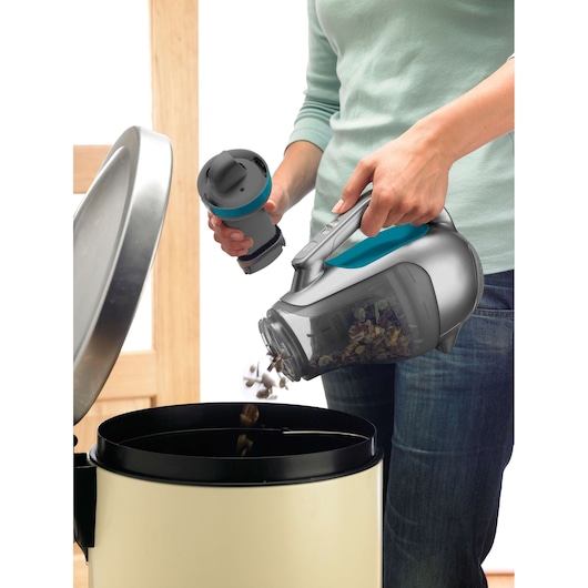 A person is emptying the contents of a handheld vacuum cleaner into a trash bin. The vacuum cleaner is gray and blue, and the person is holding its detachable dust container over the bin.