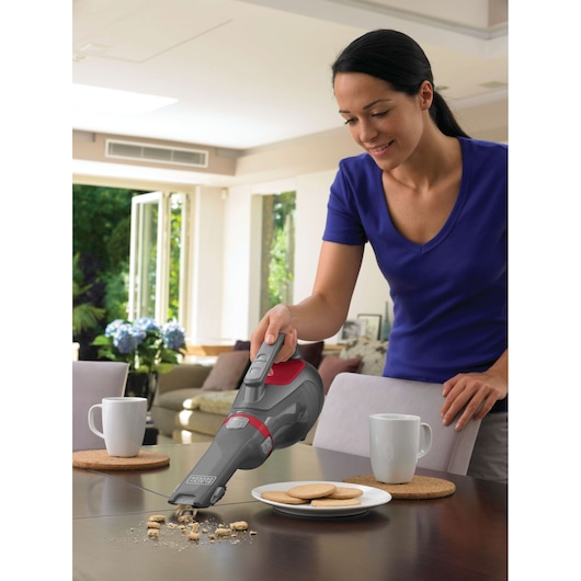 A person using a BLACK+DECKER handheld vacuum cleaner to clean crumbs from a table next to a plate of cookies and two cups in a bright living room.
