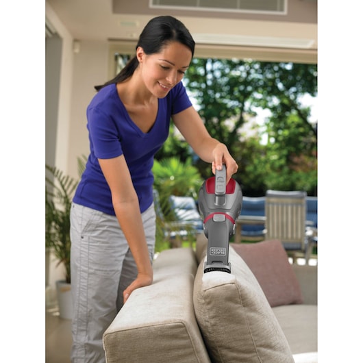 A person using a BLACK+DECKER handheld vacuum cleaner (model DV1415EL) to clean the top of a sofa in a bright indoor living area, with outdoor greenery visible through the windows.