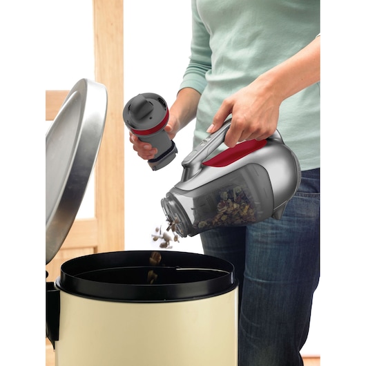 A person empties the dust container of a BLACK+DECKER handheld vacuum cleaner into a trash bin.