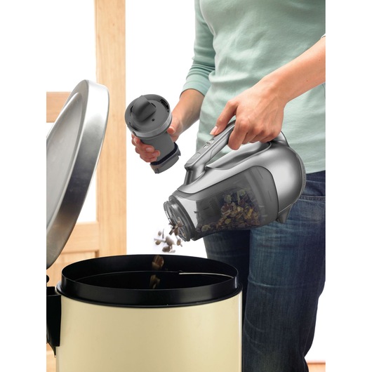 A person empties the dust container of a BLACK+DECKER handheld vacuum cleaner into a trash bin.