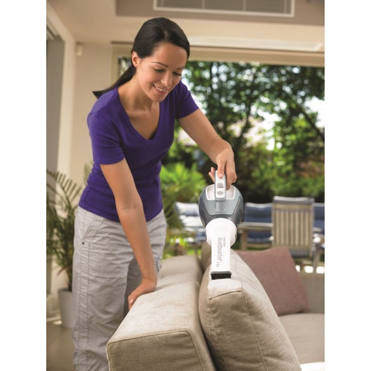 A person using a BLACK+DECKER Dustbuster handheld vacuum cleaner to clean the side of a beige sofa in a bright living room.