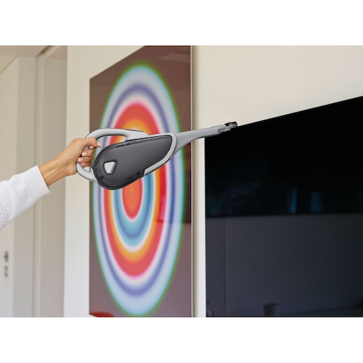 A person uses a BLACK+DECKER handheld vacuum cleaner (model DVJ215J) with a narrow nozzle attachment to clean the edge of a television screen in a modern home, with a colorful circular artwork in the background.